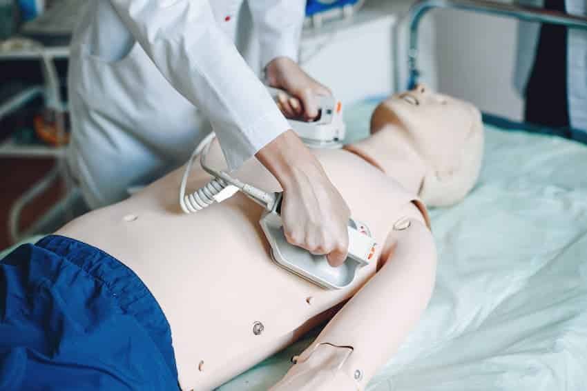 Une personne gantée pratique une compression thoracique sur un mannequin de secourisme, sous la supervision d’un formateur.