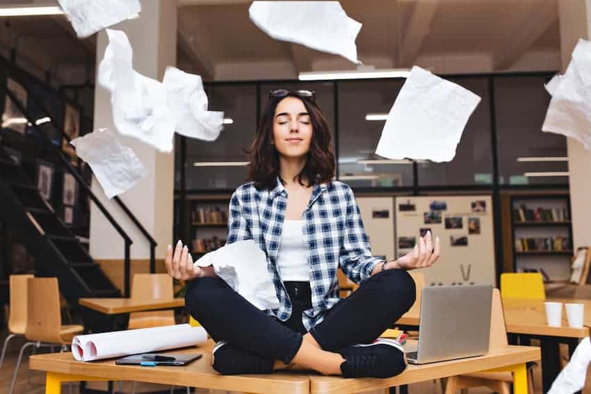 Femme méditant en position assise sur une table au milieu de papiers volants dans un bureau moderne. Elle affiche une expression calme et détendue.