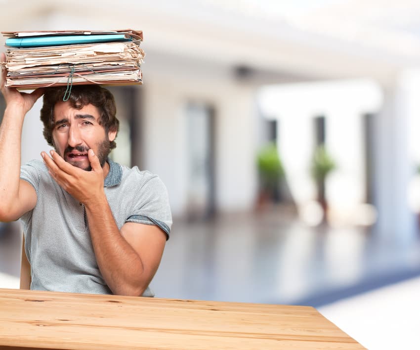Salarié débordé en entreprise équilibrant une pile de dossiers sur la tête, illustrant la surcharge de travail et les risques psychosociaux.
