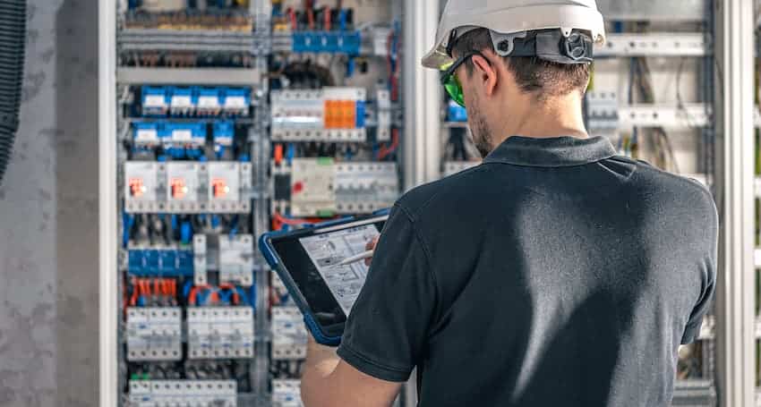 Technicien équipé d'un casque de sécurité consultant une tablette devant un tableau électrique, illustrant la prévention du risque électrique en formation H0B0.