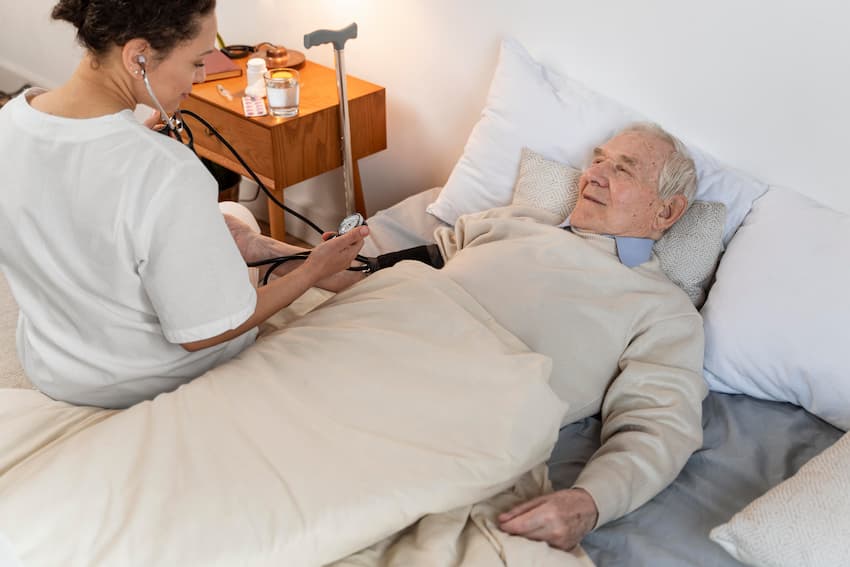 Soignante prenant la tension artérielle d'un résident âgé alité, illustrant les soins de confort et l'accompagnement bienveillant en fin de vie en EHPAD.