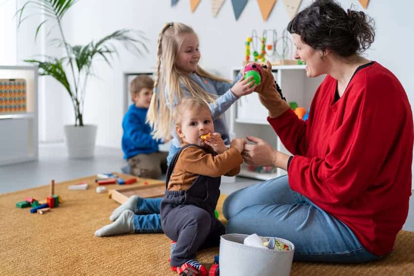 Professionnelle de la petite enfance assise au sol avec des enfants lors d'une activité d'éveil, illustrant les postures contraignantes à l'origine des risques physiques en crèche.