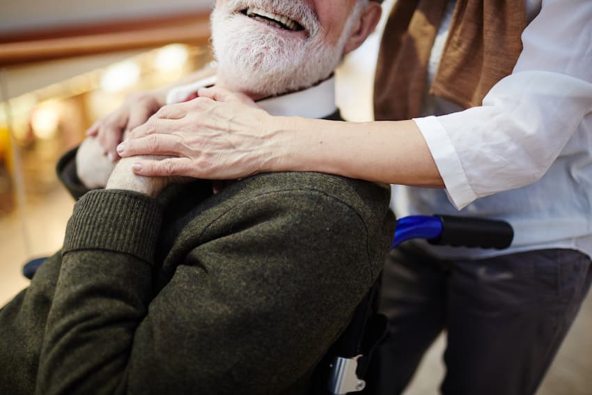 Soignante posant les mains sur les épaules d'un résident âgé en fauteuil roulant, illustrant la relation bientraitante et le respect de la dignité au cœur de la réforme Maison France Autonomie.