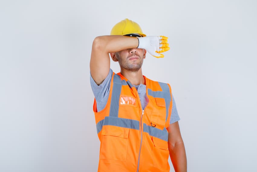 Travailleur en équipement de protection individuelle se protégeant les yeux avec son avant-bras dans un geste d'épuisement ou d'éblouissement, évoquant la fatigue et la surexposition à la chaleur ou à la lumière sur les chantiers.