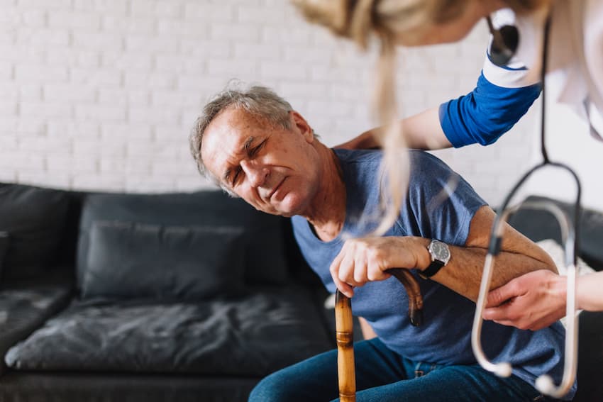 Un soignant aide un homme âgé à se lever d'un canapé, en soutenant son bras. L'homme s'appuie sur une canne et grimaçe sous l'effort.
