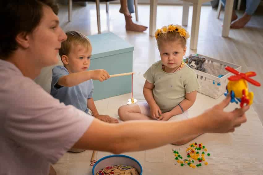 Professionnel de la petite enfance animant une activité de jeu au sol avec deux jeunes enfants, illustrant l'organisation de la formation PRAP Petite Enfance en crèche.