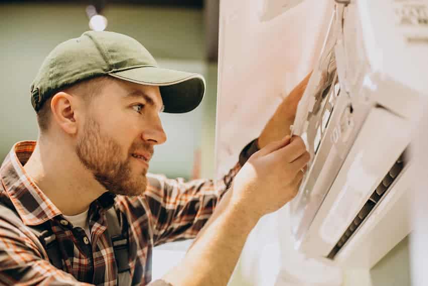 Technicien en casquette verte et chemise à carreaux intervenant sur une unité intérieure de climatisation murale, regard concentré sur l'appareil, profil gauche, arrière-plan flou d'atelier ou de local technique.
