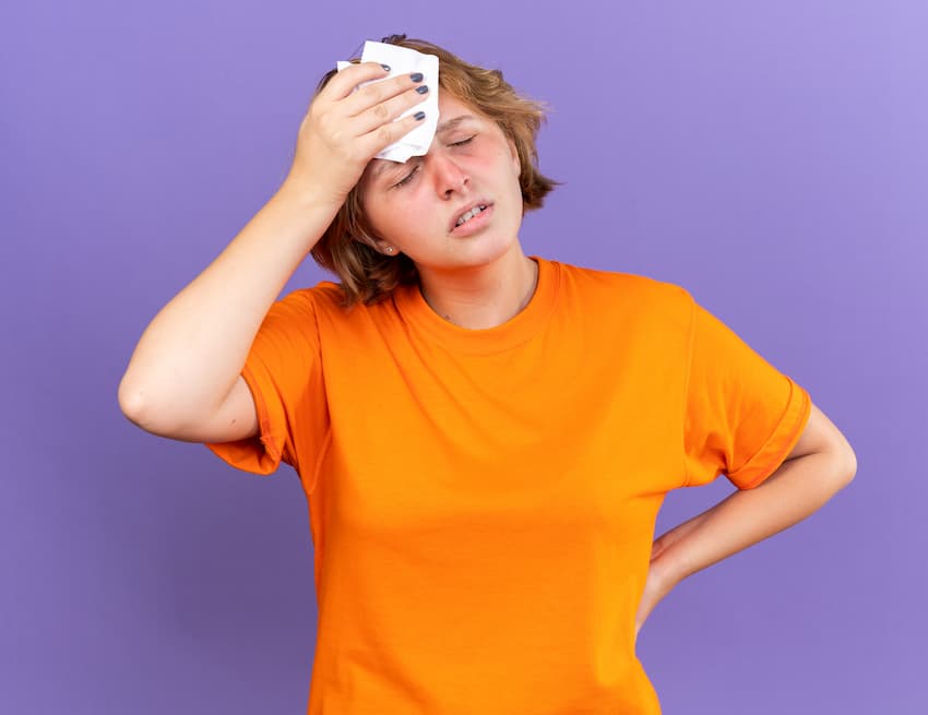 Jeune femme en t-shirt orange appliquant un tissu humide sur le front, yeux mi-clos, expression douloureuse, main gauche sur la hanche, fond violet uni, vue de face.