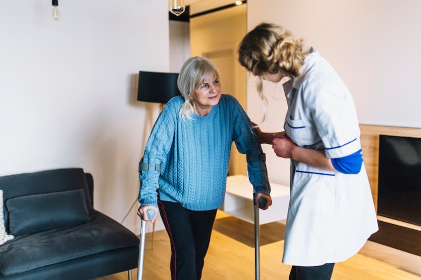Soignante accompagnant une résidente âgée marchant avec des béquilles, illustrant la mise en place d'un programme de prévention des chutes et de rééducation en établissement.