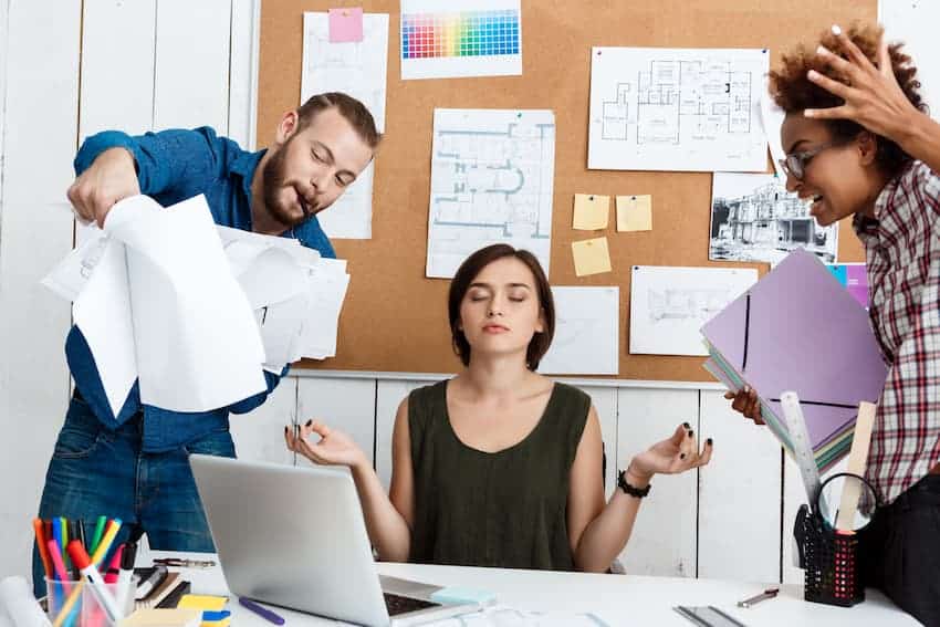 Femme méditant à son bureau pendant que deux collègues agités s'emportent autour d'elle, dans un studio de création.