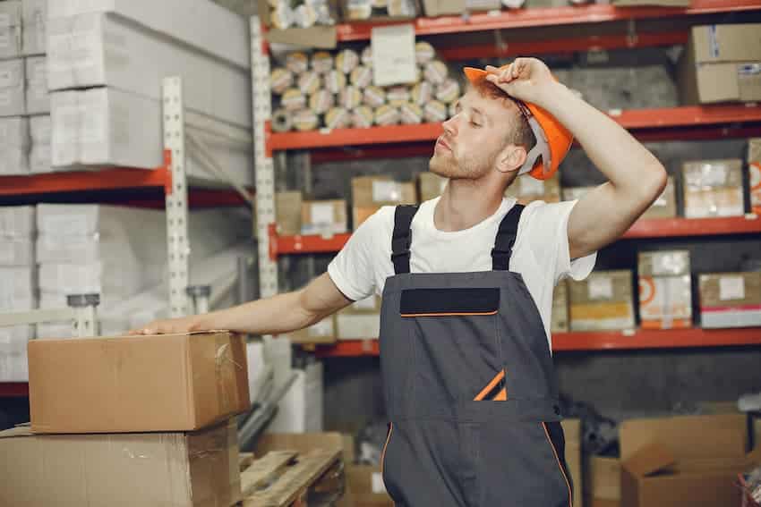 Manutentionnaire en entrepôt marquant une pause près de cartons empilés, illustrant la fatigue physique et les risques de TMS liés à la manutention et au port de charges.