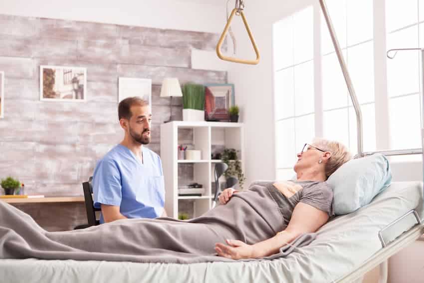 Soignant échangeant avec une résidente âgée alitée équipée d'une potence de lit, illustrant la dimension relationnelle de la mobilisation des personnes aidées dans le secteur sanitaire et social.