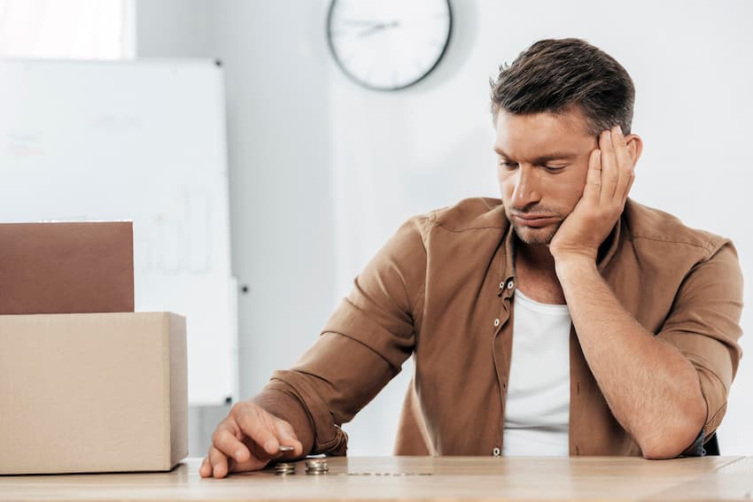 Homme résigné assis à son bureau, comptant des pièces près d'un carton de départ, dans un bureau avec horloge murale.