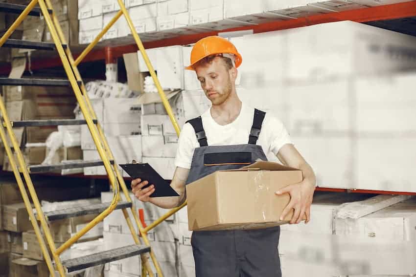 Jeune magasinier avec casque de chantier portant un carton d'une main et consultant un bon de commande dans un entrepôt.
