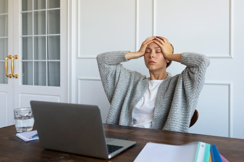 Salariée assise à son bureau, les mains sur la tête, face à son ordinateur portable et à des dossiers en attente.