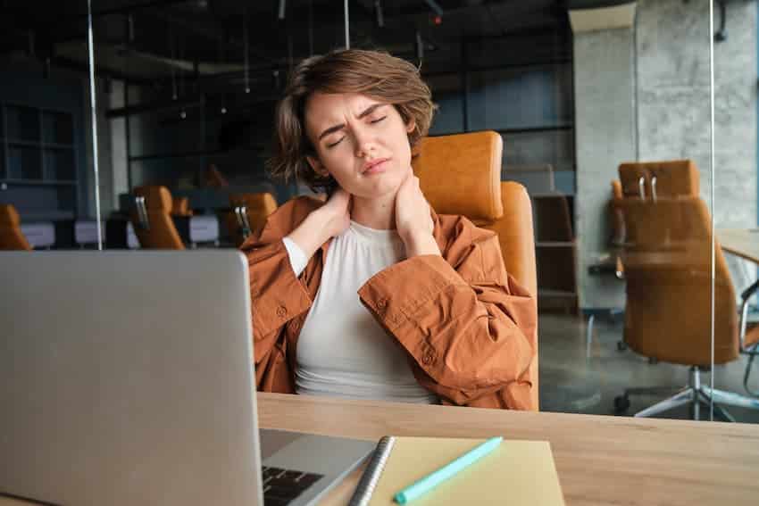 Salariée assise devant son ordinateur portable, se tenant la nuque à deux mains avec une grimace de douleur.